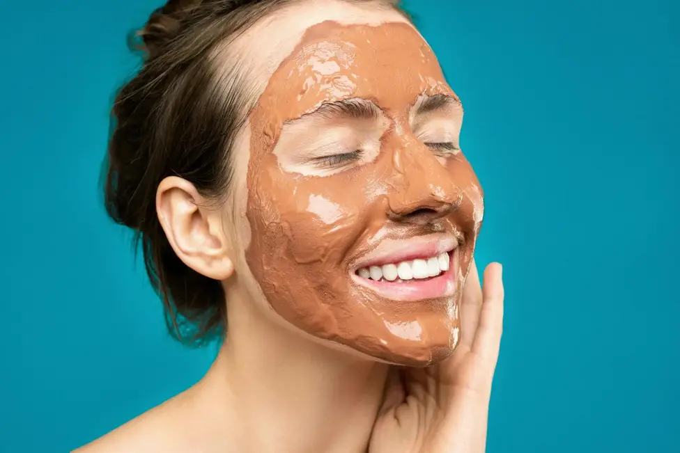 close up of a woman smiling with a clay mud mask on her face on a turquoise background GhUG6LschZUEL3 Bl6IXFg1 bk5kEMSbGczuS1cTfwA7A